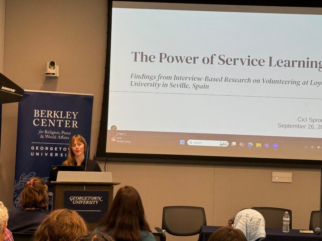 Photo of Cici Sprouse at a podium next to a screen with the slide, "The Power of Service Learning. Findings from Interview-Based Research on Volunteering at Loyola University in Seville, Spain."