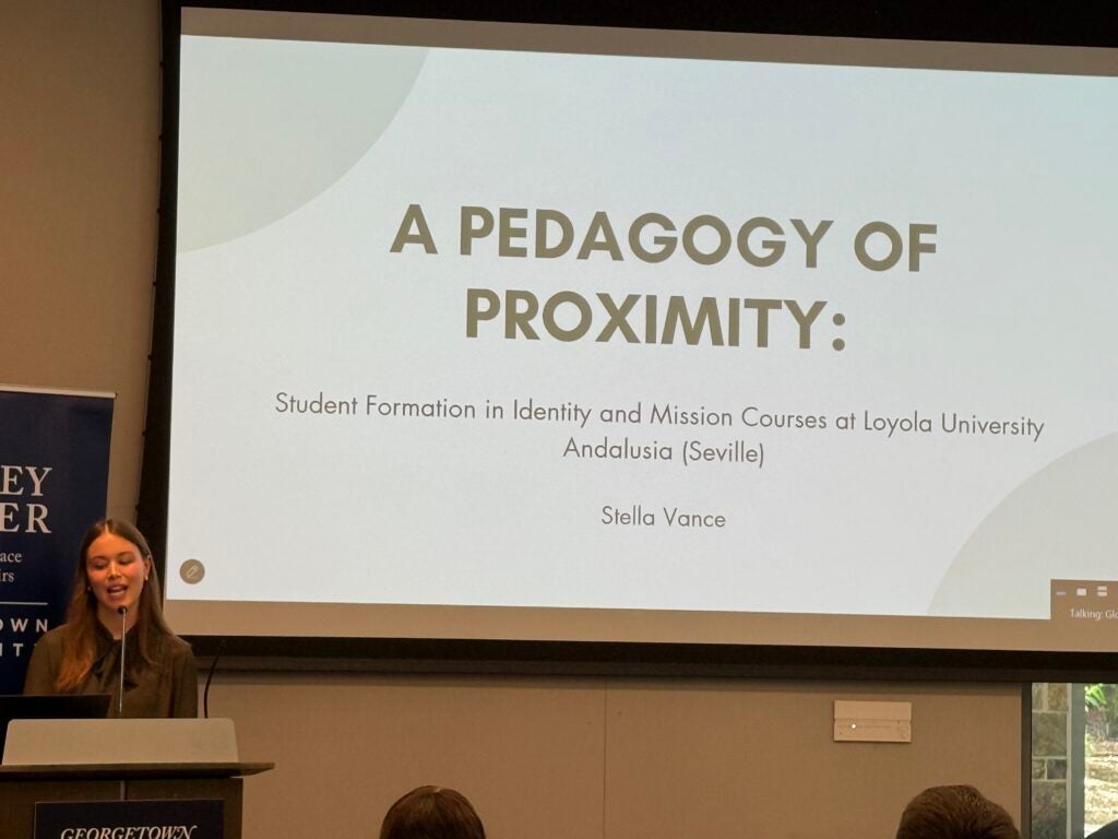 Stella Vance at a podium with a screen displaying a slide with text, "A Pedagogy of Proximity: Student Formation in Identity and Mission Courses at Loyola University Andalusia (Seville)"